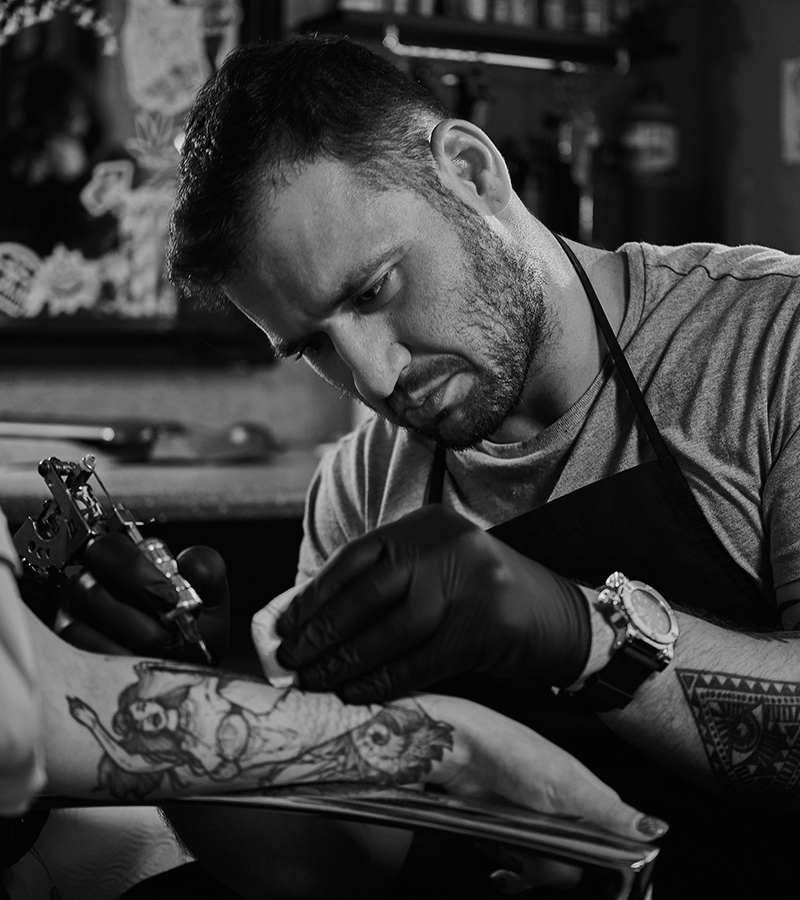 Un tatuador con barba y expresión concentrada limpia el brazo de un cliente mientras sostiene una máquina de tatuar. El cliente tiene un tatuaje en proceso con un diseño detallado.